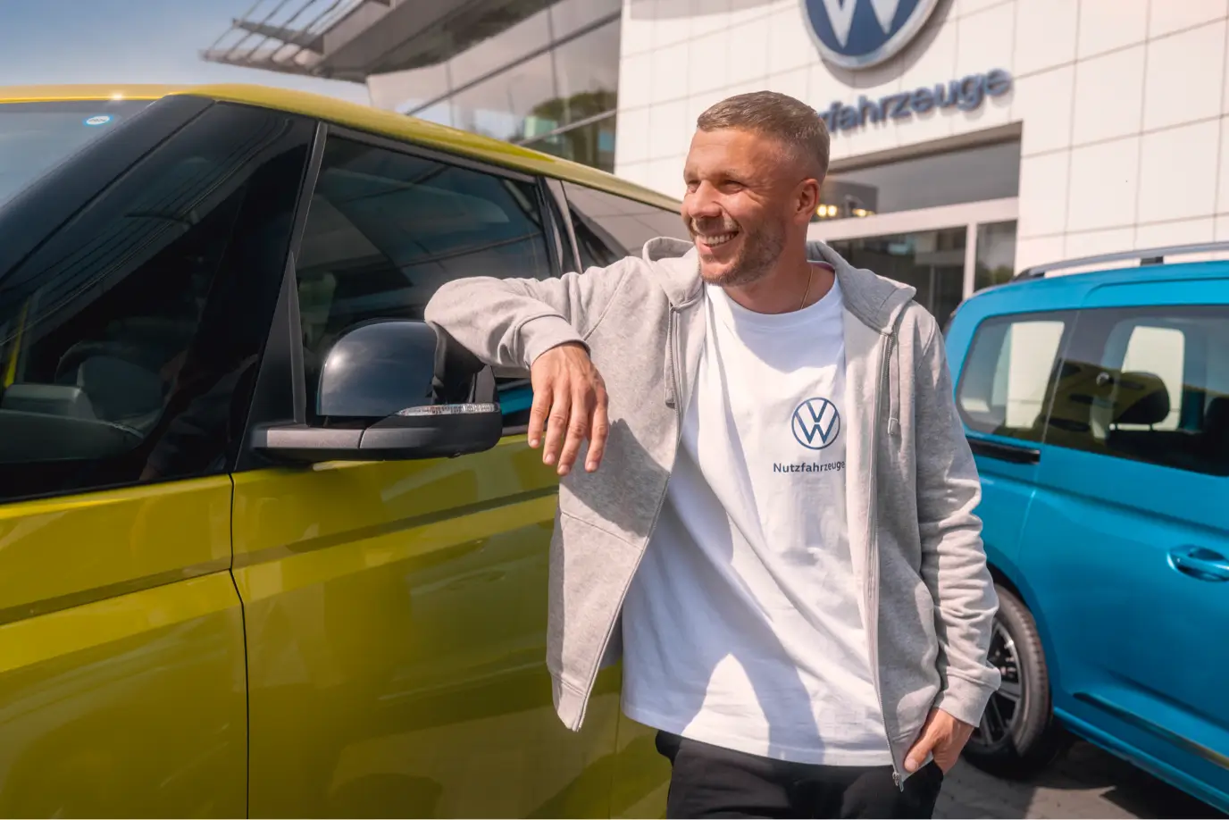 Lukas Podolski lehnt lachend an einem gelben Volkswagen ID. Buzz vor einem VW Nutzfahrzeuge Autohaus, daneben ein blauer Caddy.